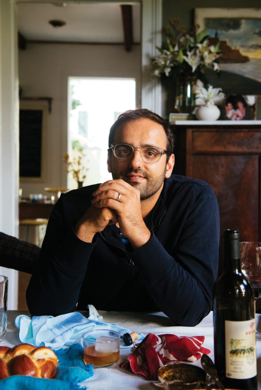 Photo of Alon Shaya smiling at a table with a bottle of wine and a loaf of challah bread. 