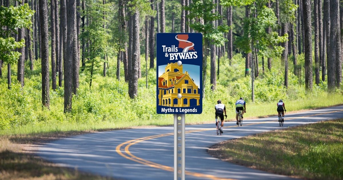 Bikers on road surrounded by woods with a sign saying "Trail & Byways--Myth & Legends" in Vernon Parish for a Louisiana road trip