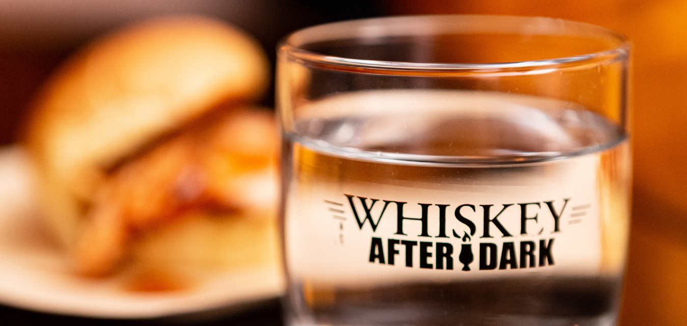 Photograph of a glass that says Whiskey After Dark and a BBQ sandwich in the background