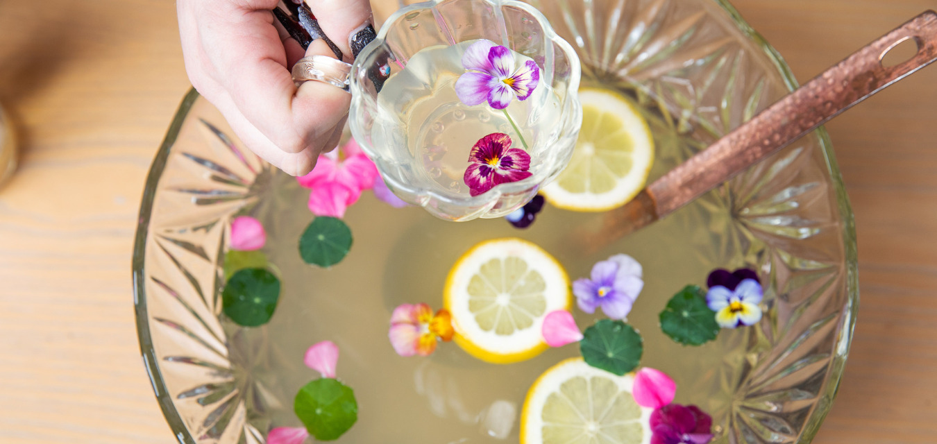 A glass punch bowl with a yellow-ish liquid and pink flowers and lemon slices inside it.