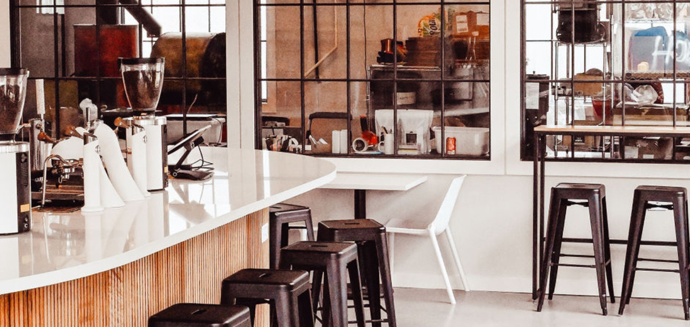 The Interior of Progress Coffee. A white counter and walls, dark brown stools at the counter, windows showing the back of the coffee shop. Minimalist style.