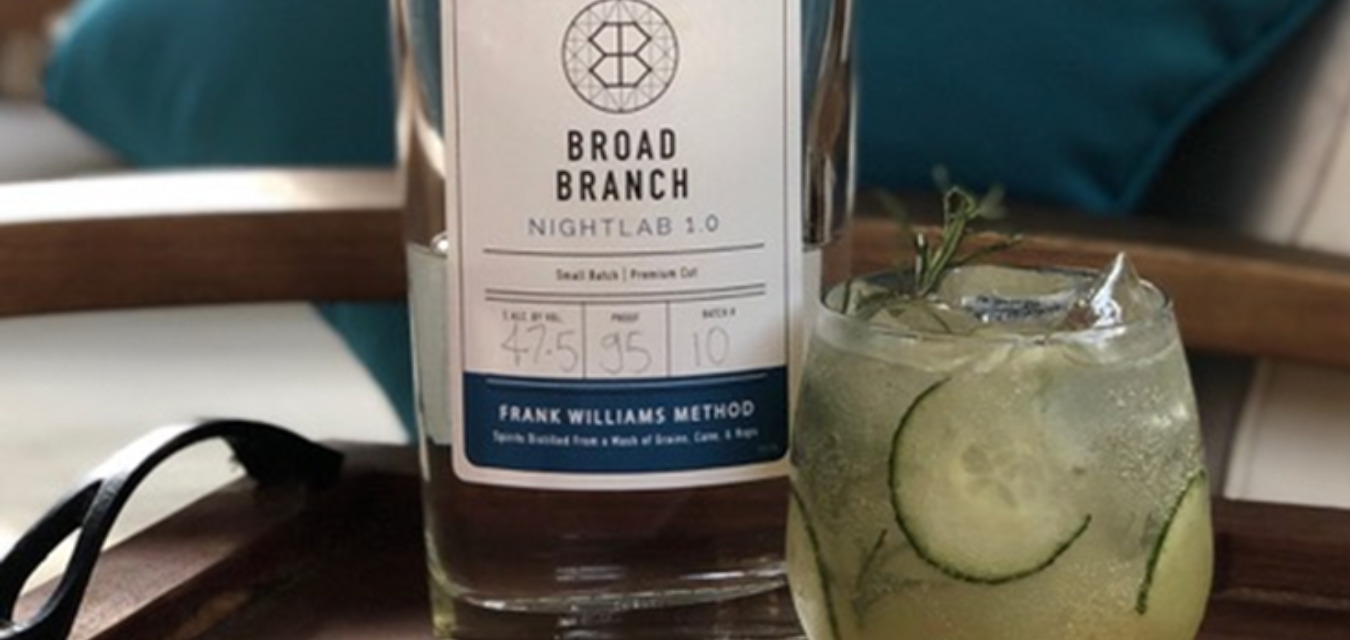 A clear bottle of Broad Branch Nightlab 1.0 on a wooden tray next to a glass of the Cocktail: Winston Collins which has a yellow-ish tint and slices of cucumber and a sprig of rosemary inside.