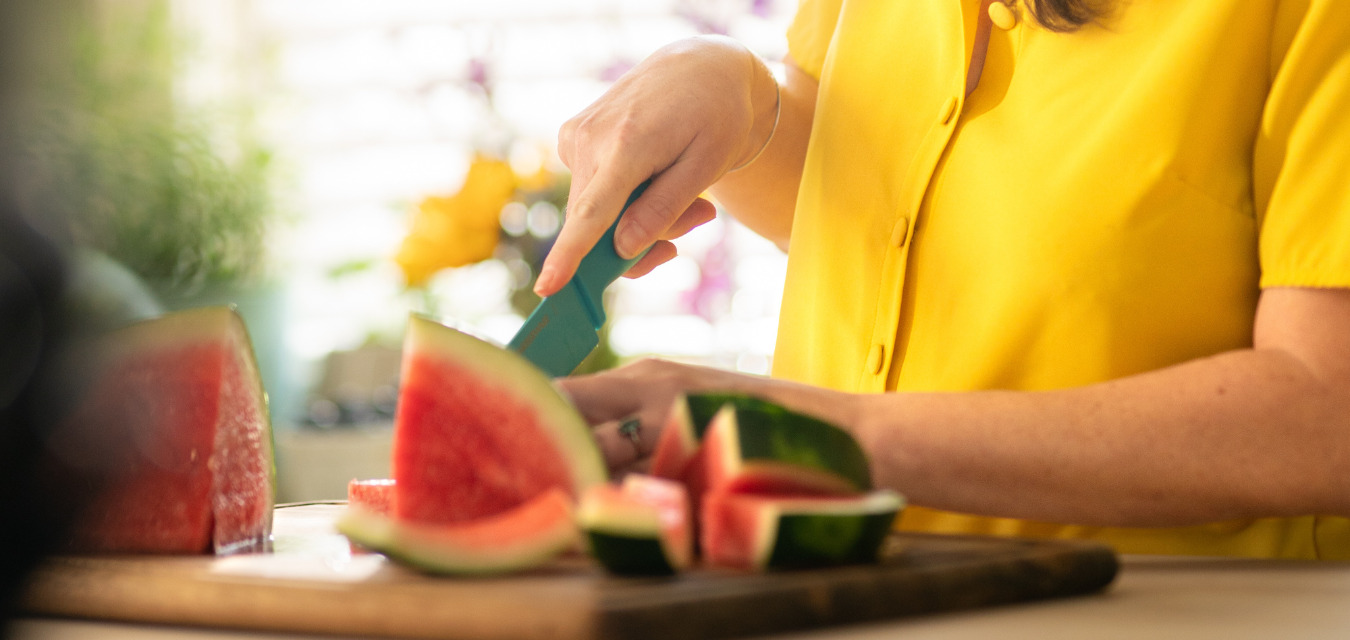 Sunlit kitchen with a woman in a bright yellow shirt. She is slicing watermelon.