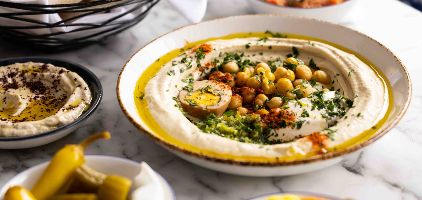 Hamsa Baba Ganoush in a white bowl topped with chickpeas, herbs, and an egg