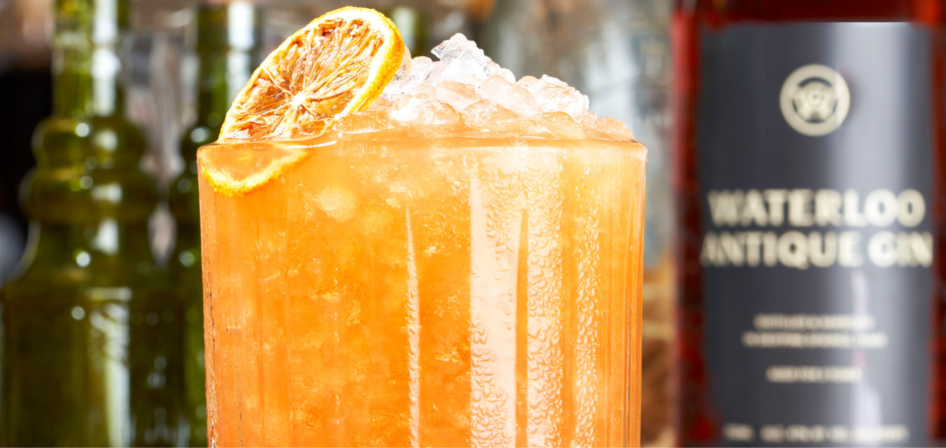 A glass of bright orange liquid with a slice of dehydrated lemon and lots of ice. A Rhubarbra Streisand, a low alcohol cocktail.