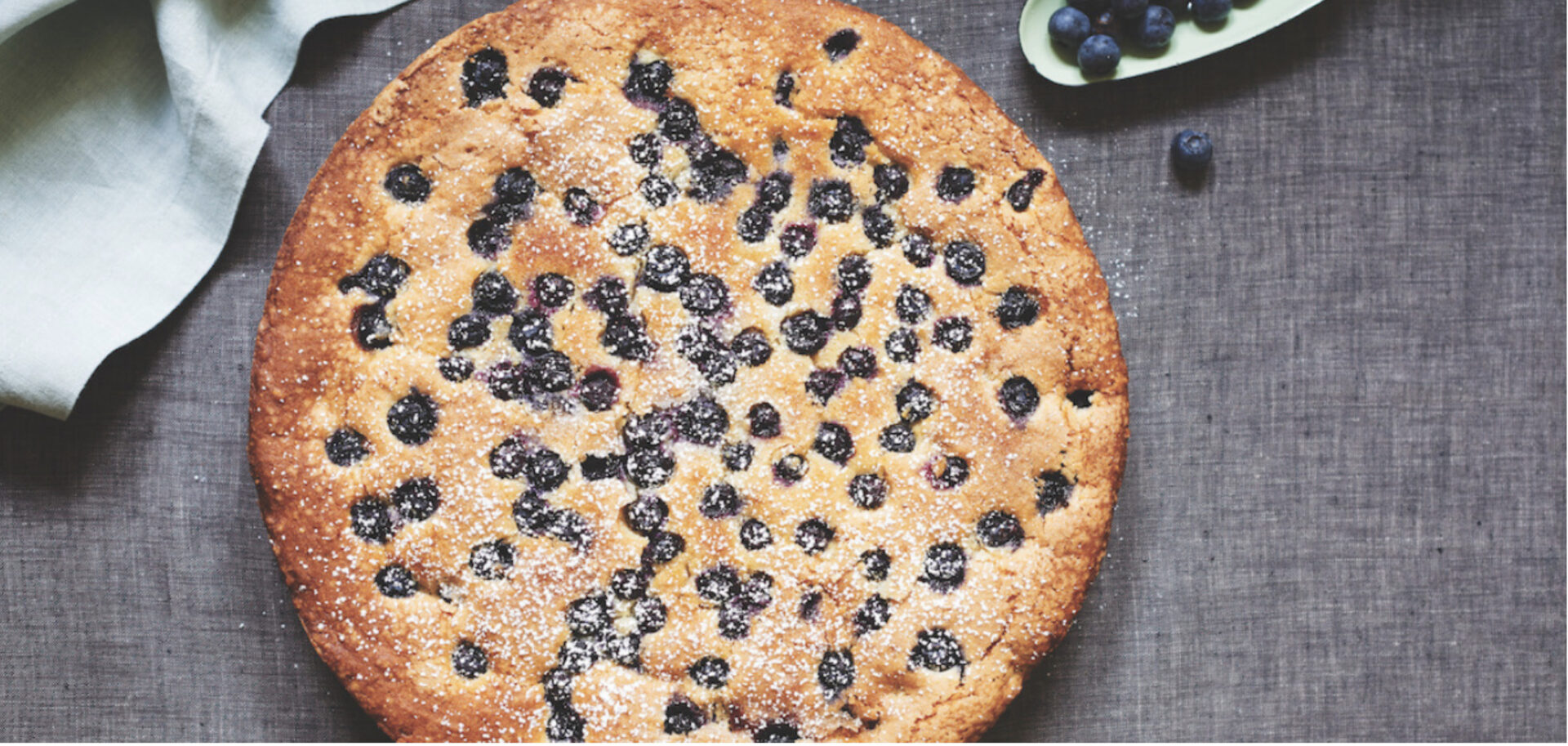 Blueberry Lemon Ricotta cake on a grey background with a napkin and stray berries beside it