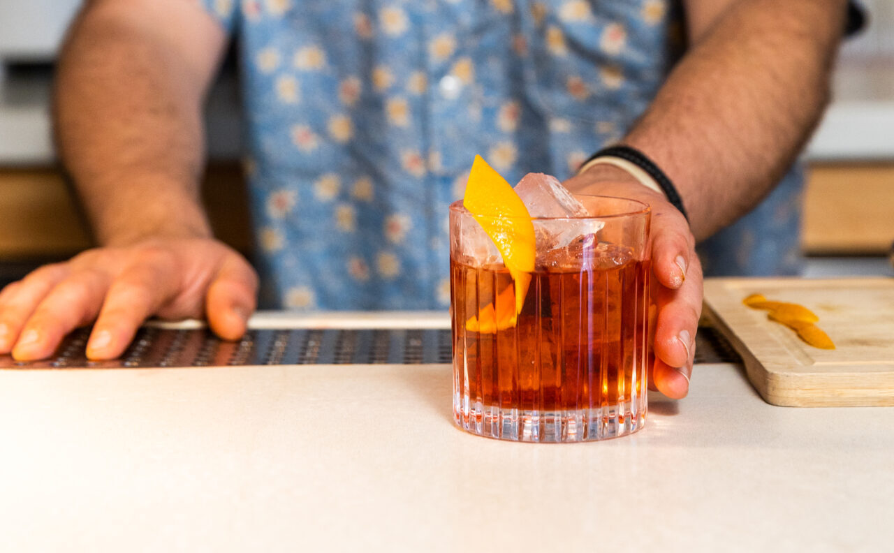 A man in a blue shirt behind a bar pushes a negroni with an orange twist into view.