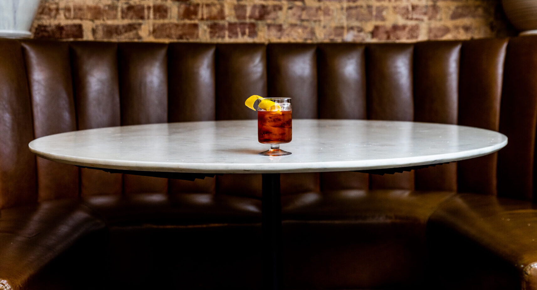 A fluted glass holds a negroni with a lemon twist on top of a white table in front of a brown booth.
