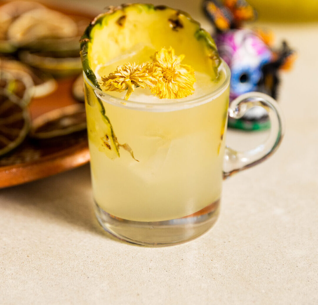 A large glass bowl holds a yellow punch garnished with pineapple, dried fruit, and flowers.
