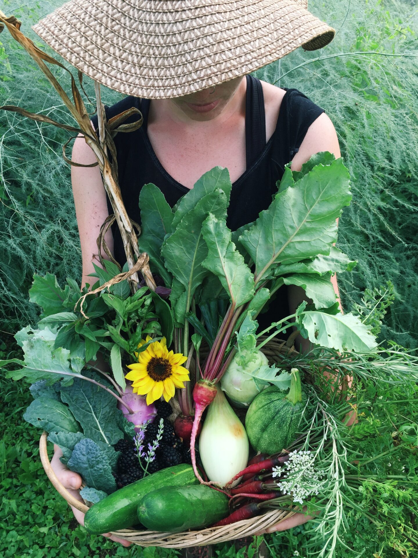 Woman holds a basket of vegetables and flowers while wearing a straw hat on the grounds of Patomack Farm 