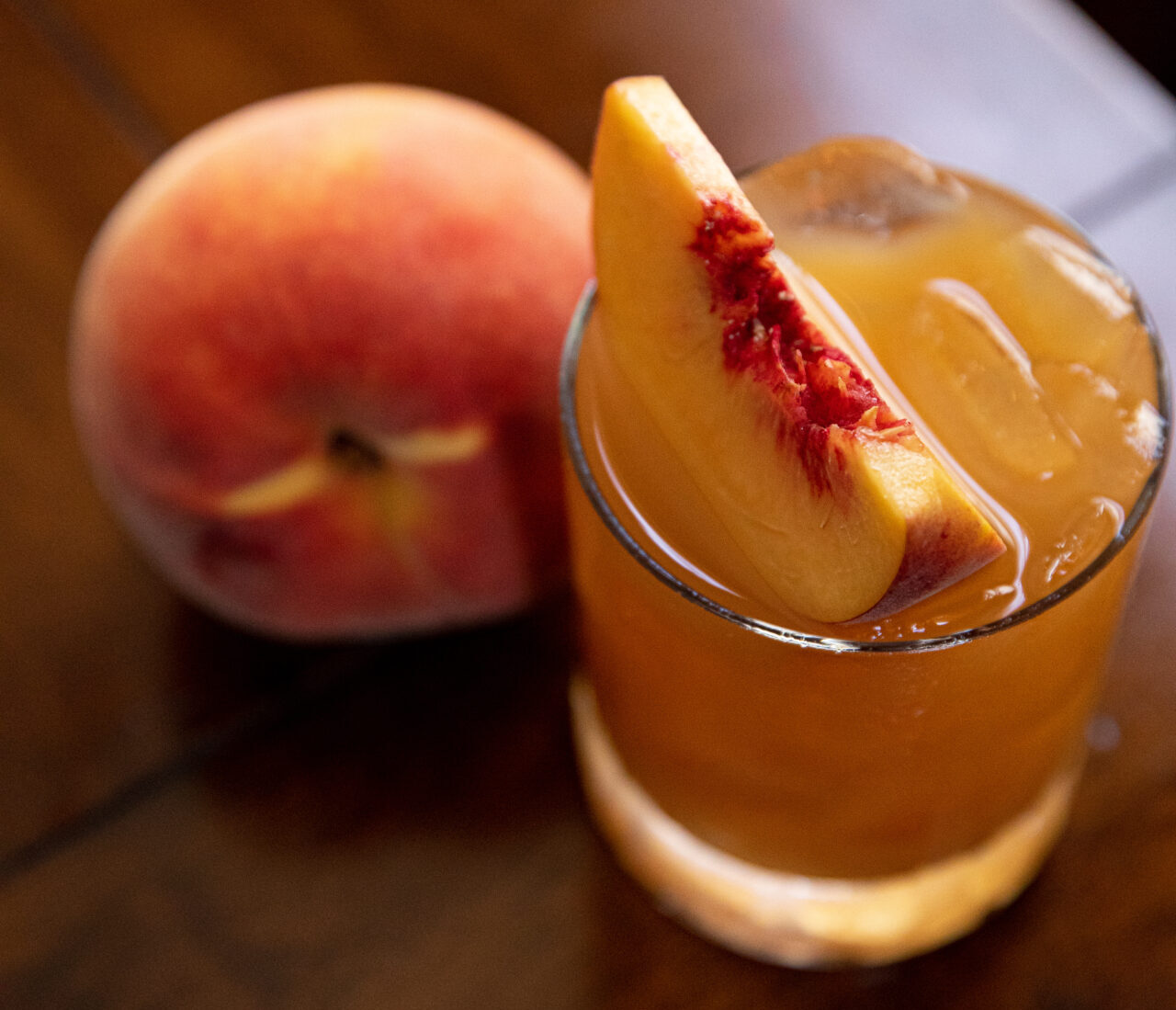 A glass of spirits sits by a peach on a table.