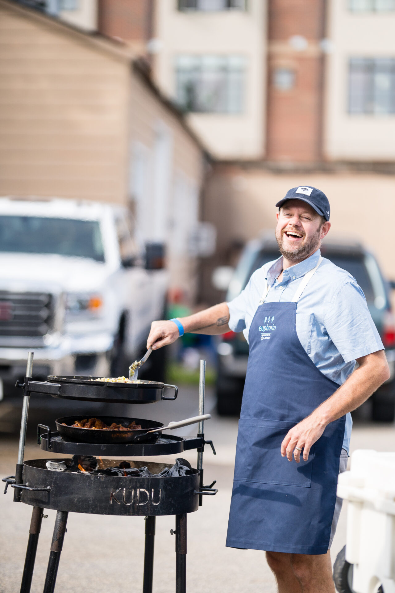 A man in a blue apron stands laughing while he grills.