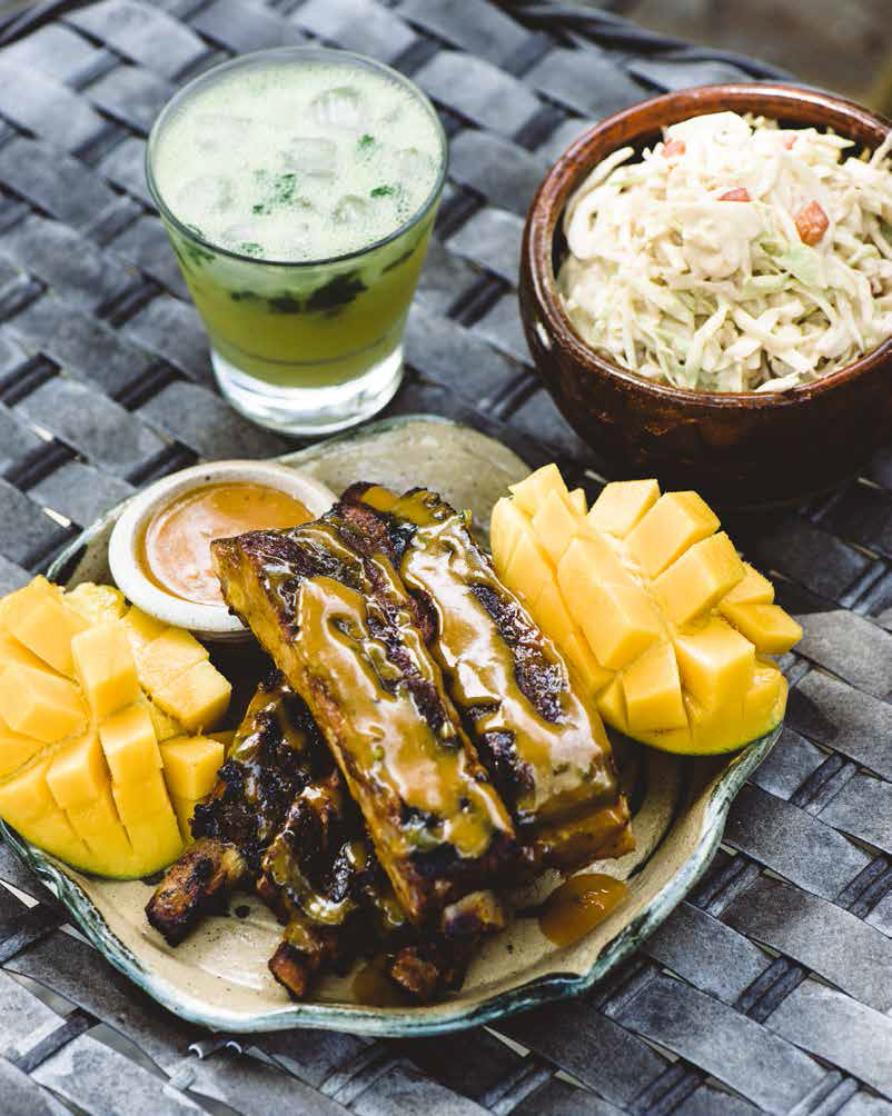 A plate of ribs, mango, a cocktail, and coleslaw sits on a table.