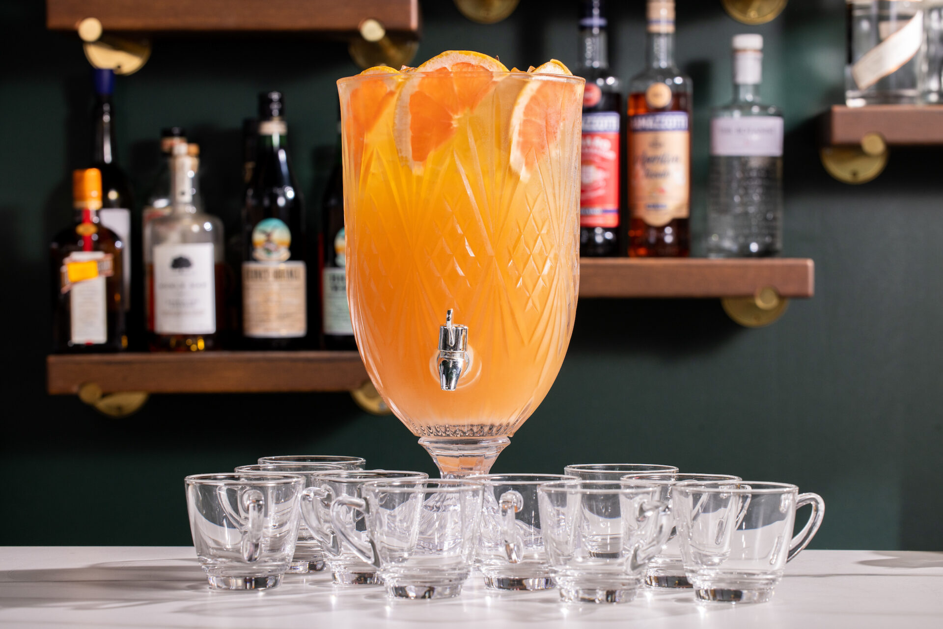 Cocktails: A pitcher sitting on a table with an orange colored punch topped with grapefruit slices. Mini glasses for pouring sit in front of the pitcher.