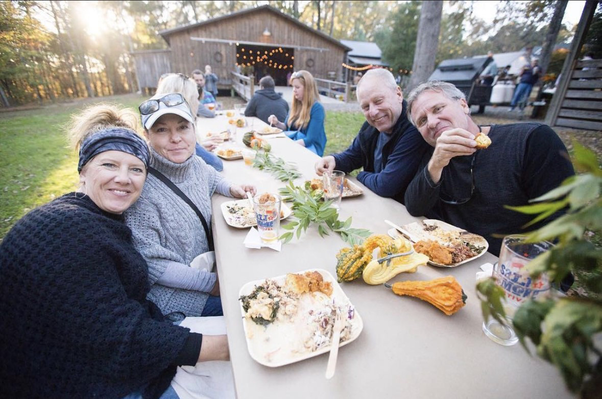 Salt & Smoke festival-goers sitting at an outdoor picnic table smiling at the camera while enjoying plates of delicious BBQ and great company!