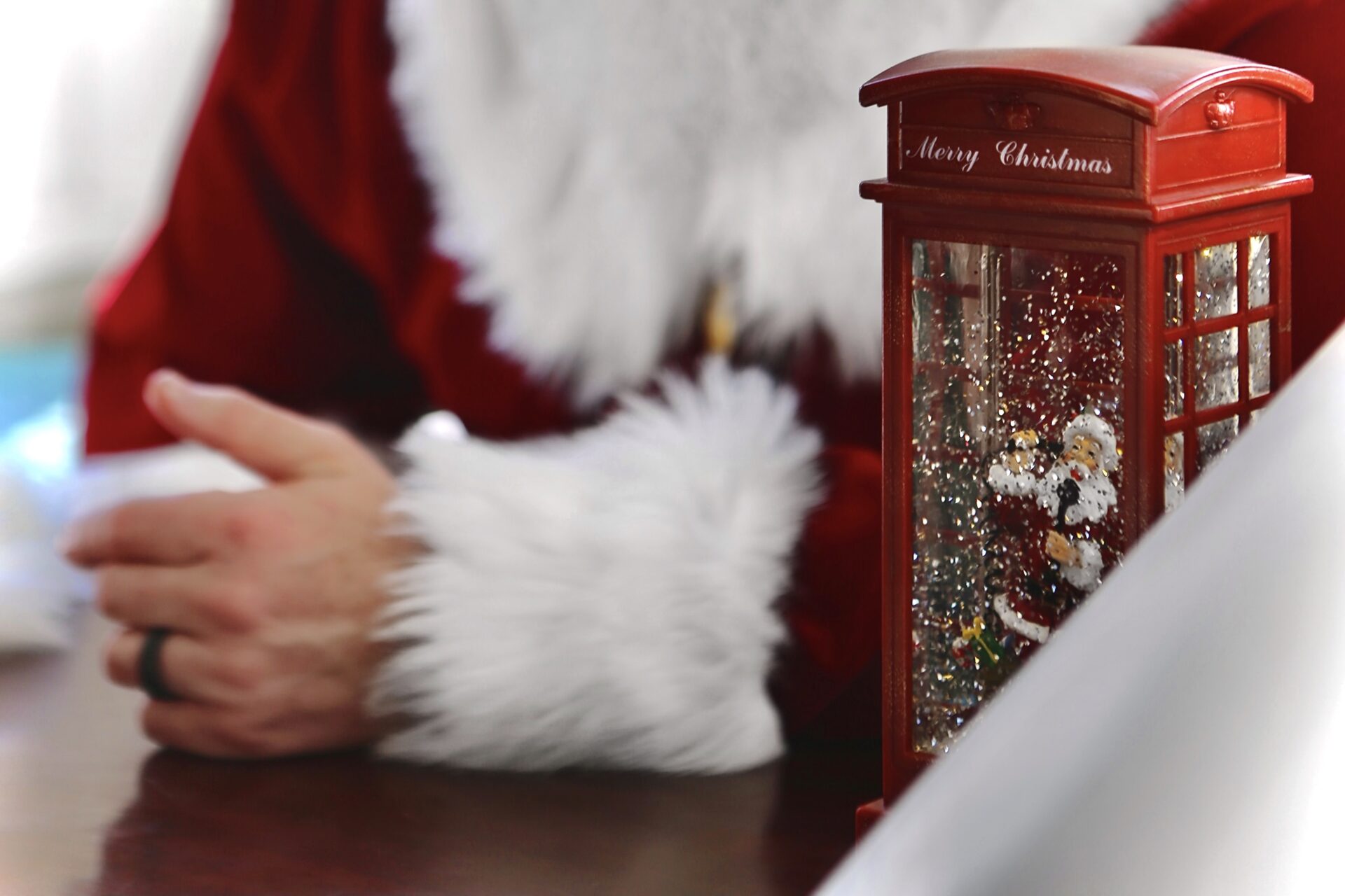 A photo of a small phone booth with a Santa action figure inside sitting on a table in front of someone dresses as Santa. 