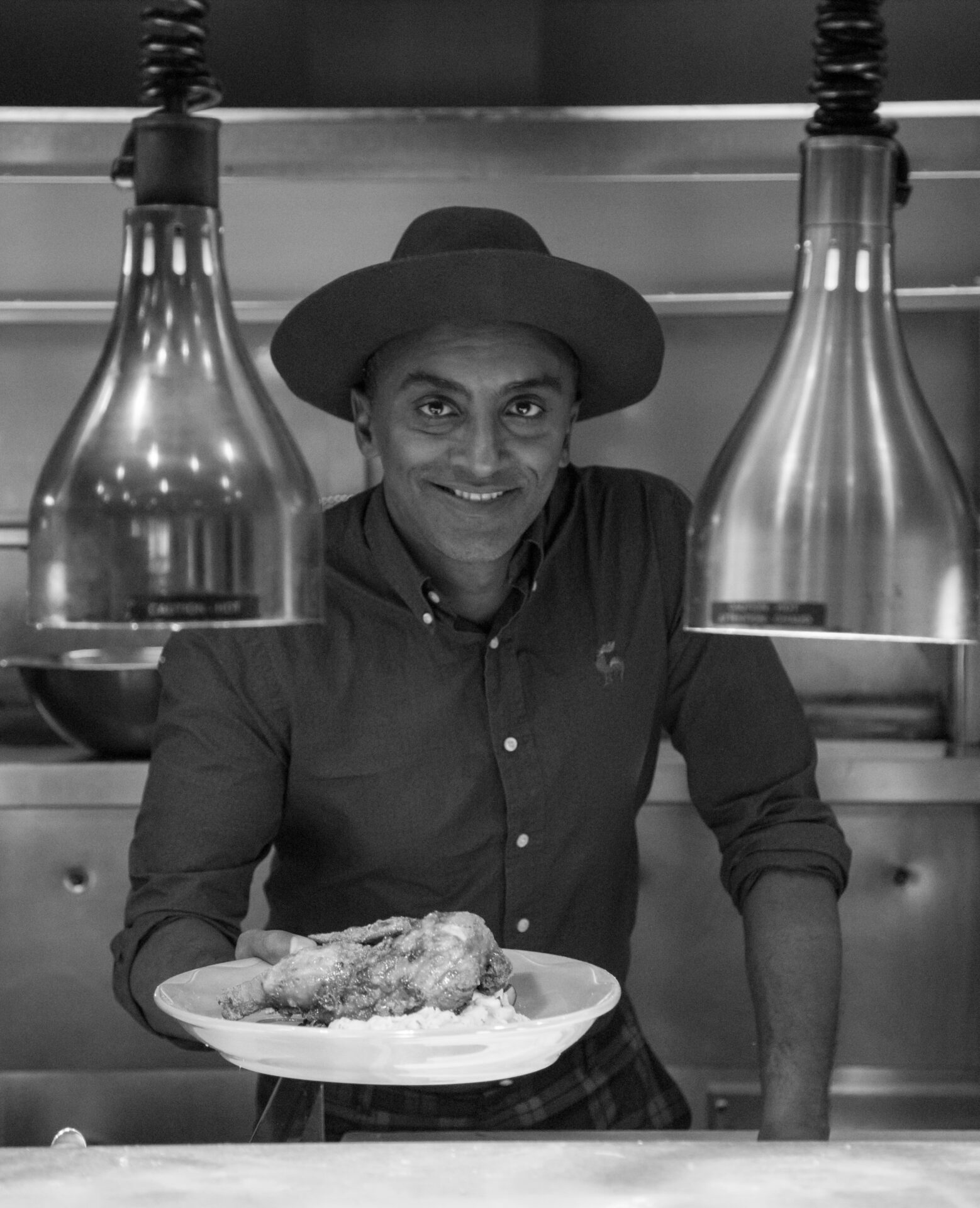In The Fridge: Black and white photo of Chef Marcus Samuelsson holding out a plate of food in a white bowl behind the line in his kitchen.