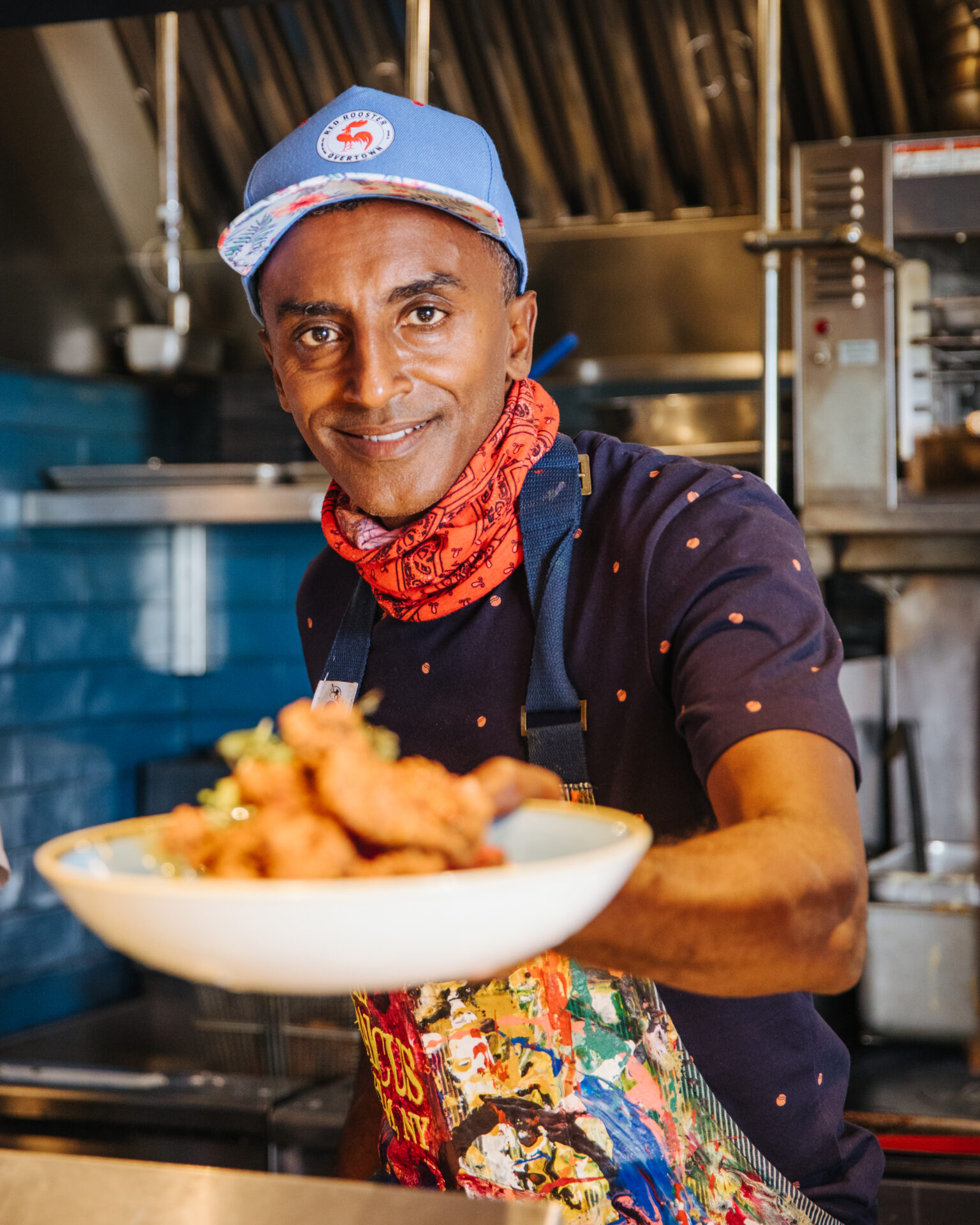 In The Fridge: Chef Marcus Samuelsson holding out a plate of food in a white bowl while standing in his colorful kitchen.