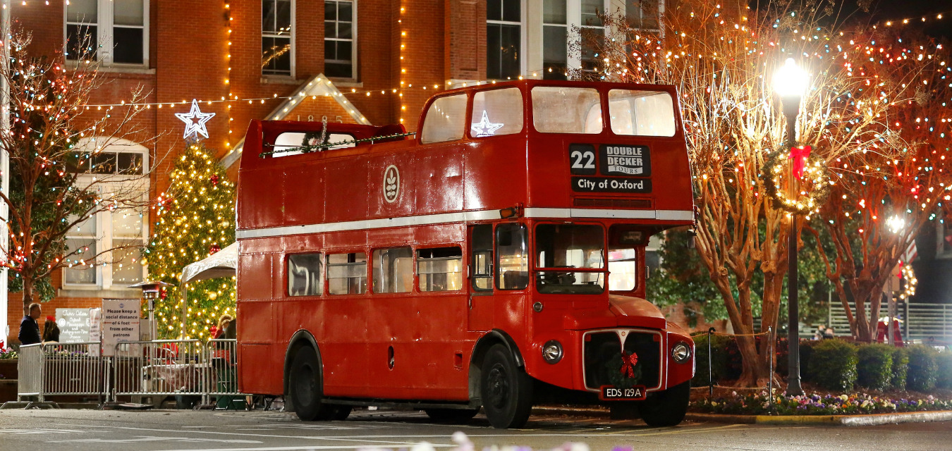 Image of a red double decker bus in front of houses decorated for the holidays.