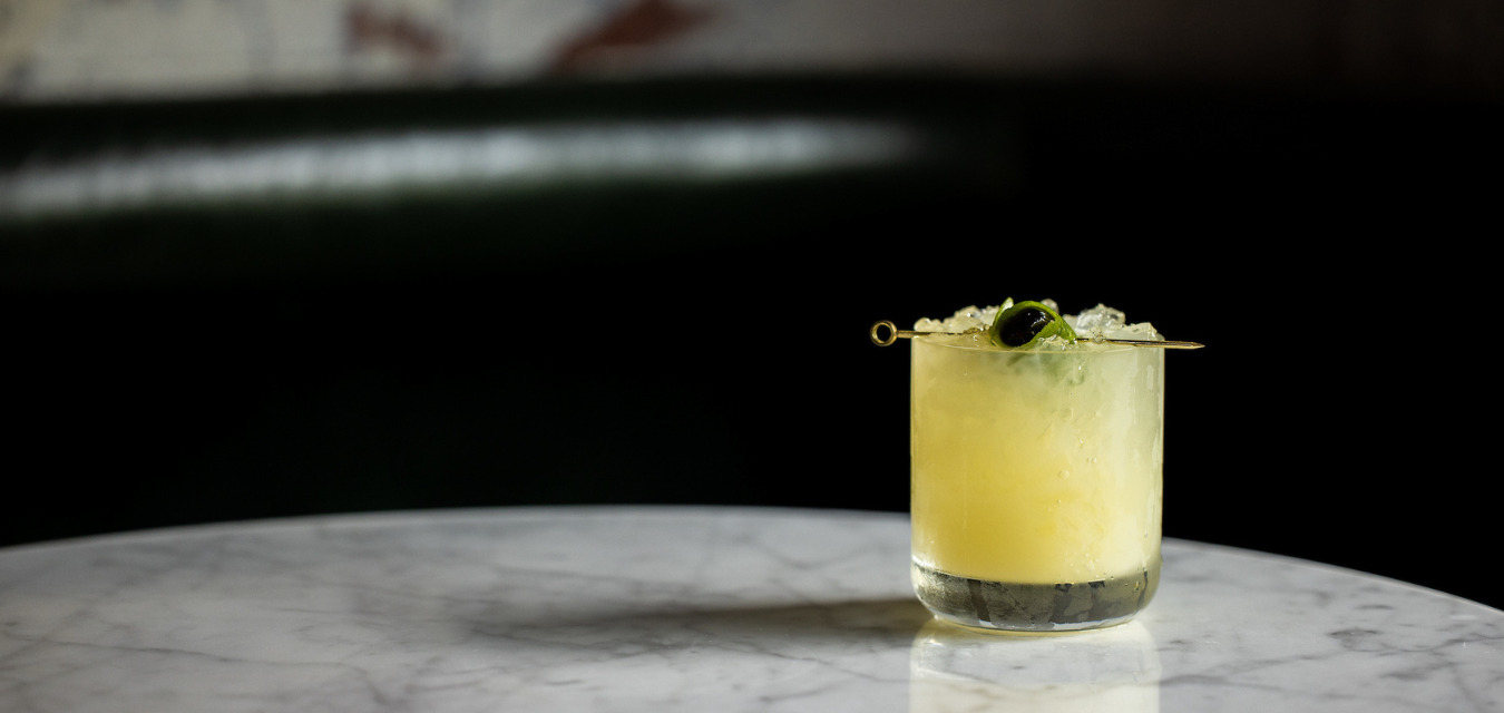 The Saturn: A glass on a table filled with a bright yellow cocktail topped with a lime and black cherry.
