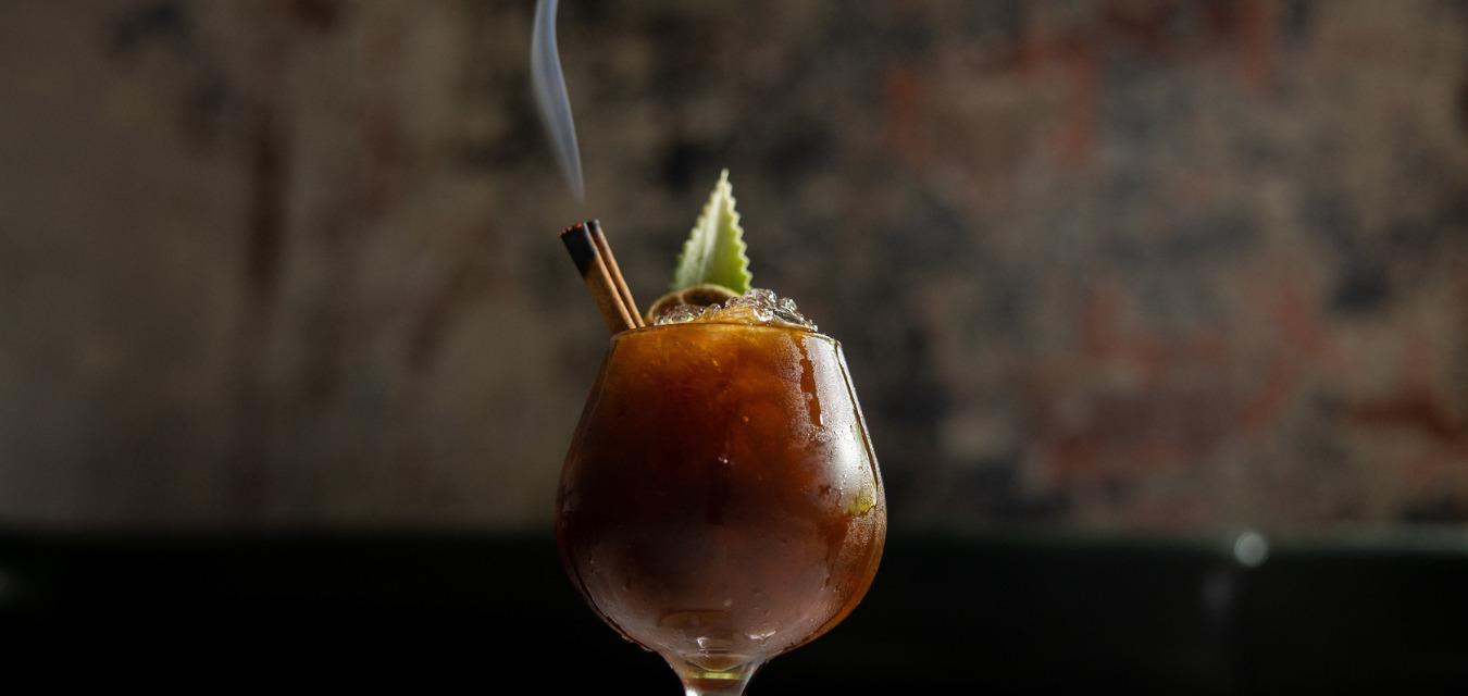 My Venus Is In Pisces: A glass sitting on a table with a brown colored drink (a sherry, chai cocktail) topped with a smoking charred cinnamon stick and a pineapple frond.