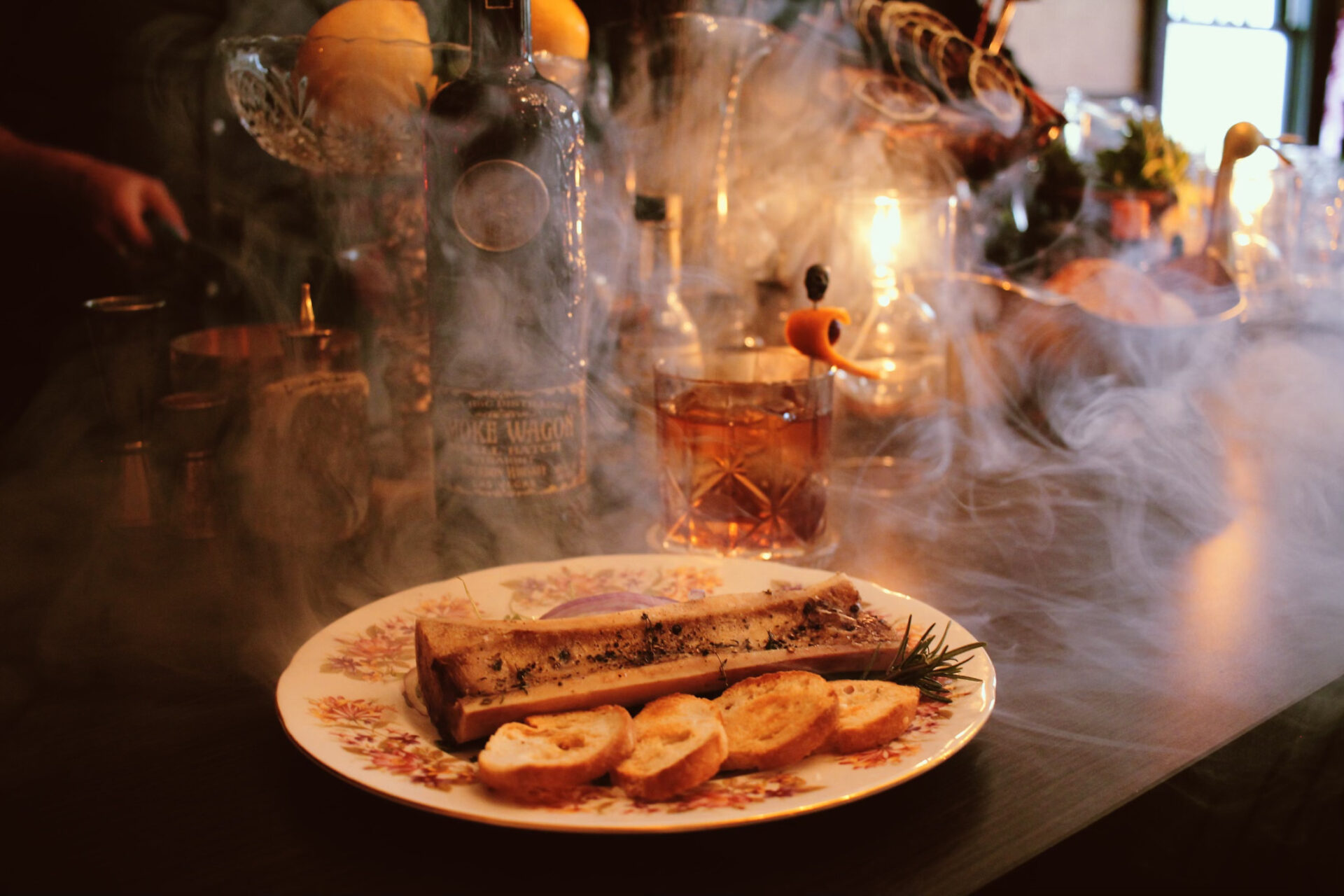 A smoky, moody dining scene with plated bone marrow, baguette slices, and a glass of whiskey at Hunter House & Gardens in Huntersville, North Carolina