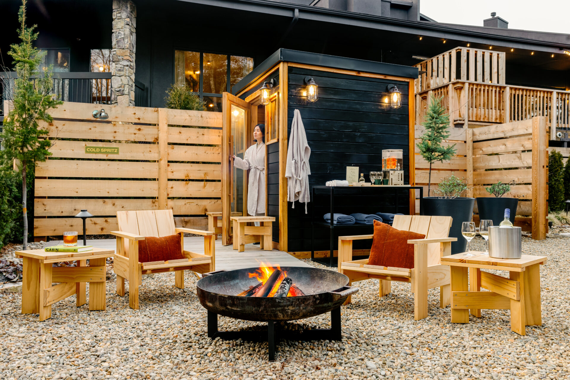 Woman in a robe exits a modern black sauna cabin at Trailborn Highlands Nordic Spa, surrounded by wooden chairs, a fire pit, and drinks on side tables.