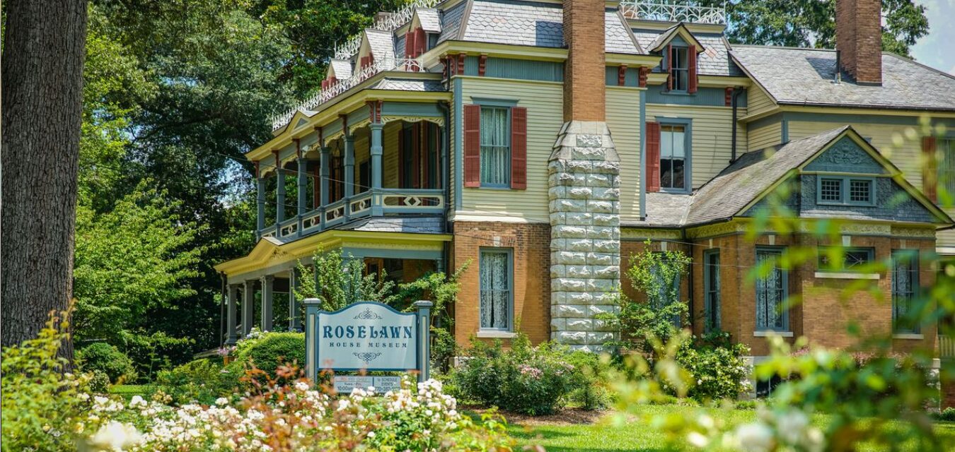 The Rose Lawn House Museum, seen from the side in this image, is a large, three-story house with multiple chimneys, two porches, lots of detail in the wood and metalworking, and painted in light, but warm shades of burgundy, green, blue, and brown. It is located in Cartersville, Georgia, known as Museum City.
