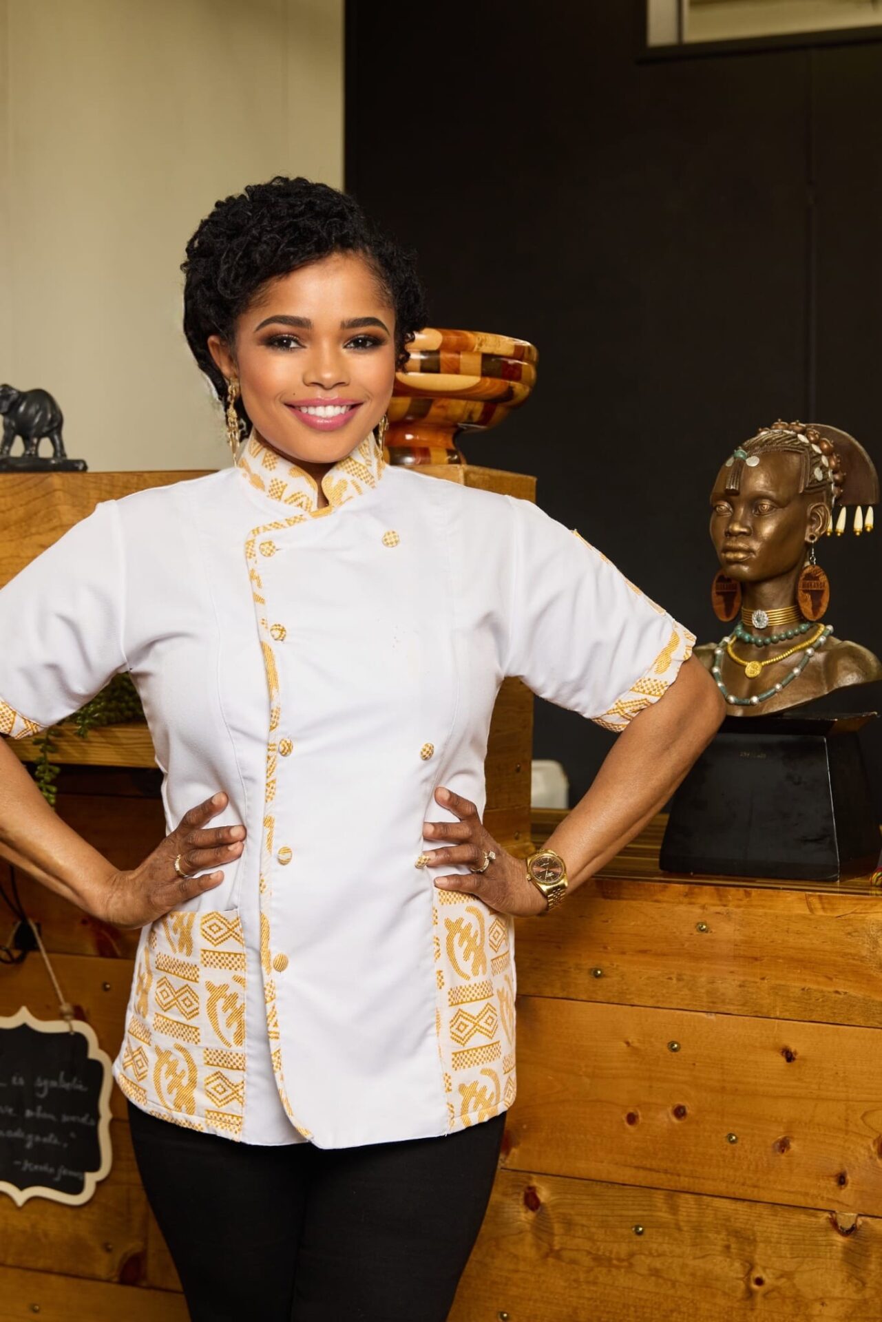 Chef Niema DiGrazia, Sierra Leonian chef in Shreveport-Bossier, Louisiana, stands confidently in a white uniform with gold details, gold jewelry, and warm makeup. 