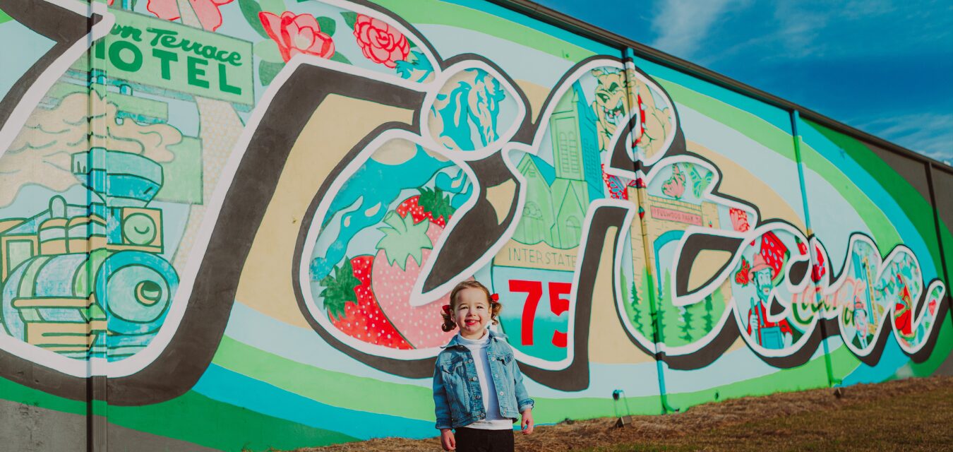 A young girl standing in front of a colorful "Tifton" mural.