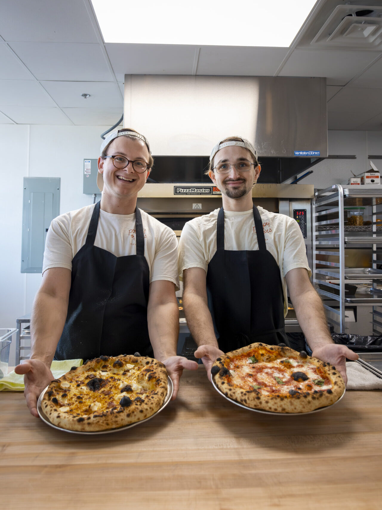 A guys wearing aprons and holding up two pizzas in the kitchen at Poppy Pies. 