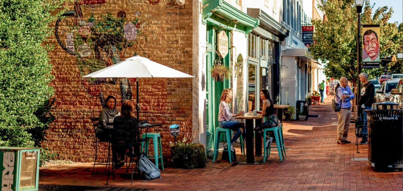 Three pairs of friends and neighbors in casual clothes chat in front of a charming, colorful, brick storefront with a mural in Lake Norman, North Carolina.