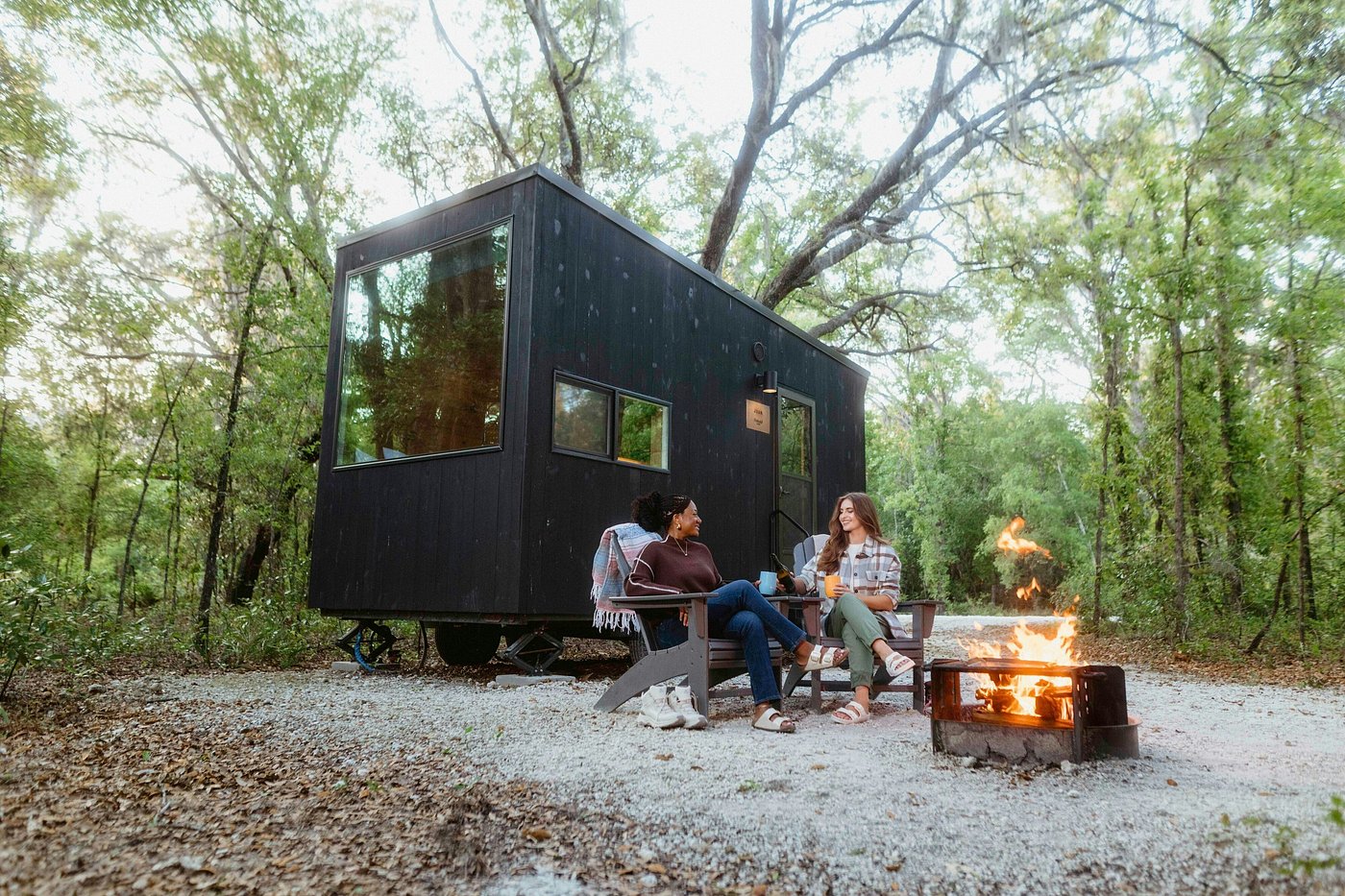 Postcard Cabins in Asheboro, a town in Randolph County, North Carolina, offer an elevated camping experience with modern, well-lit, cozy cabins. Two women enjoy a campfire outside their cabin.